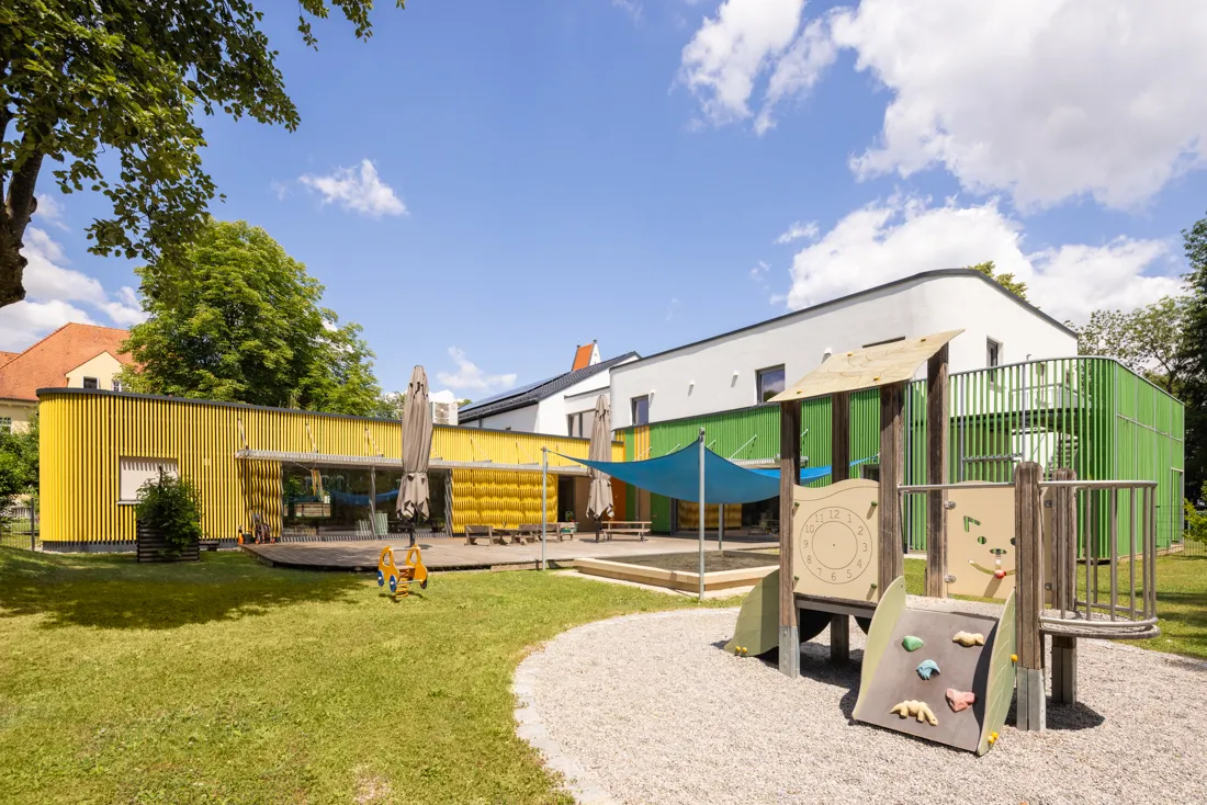 Spielplatz mit einem Baumhaus und einer Rutsche, umgeben von bunten Spielger&auml;ten und der Kindertageseinrichtung St. Mauritius im Hintergrund