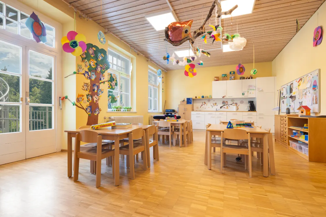 Bunter Gruppenraum mit vielen Basteleien an W&auml;nden und Decken und vielen Tischen und St&uuml;heln zum Spielen, Malen und Lernen