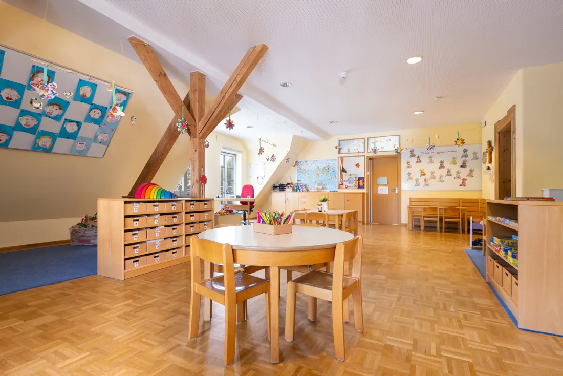 Gro&szlig;er Gruppenraum mit Dachschr&auml;ge und vielen Tischen zum Malen, Spielbereich auf dem Boden und vielen Regalen voller Spiele und Bastelsachen