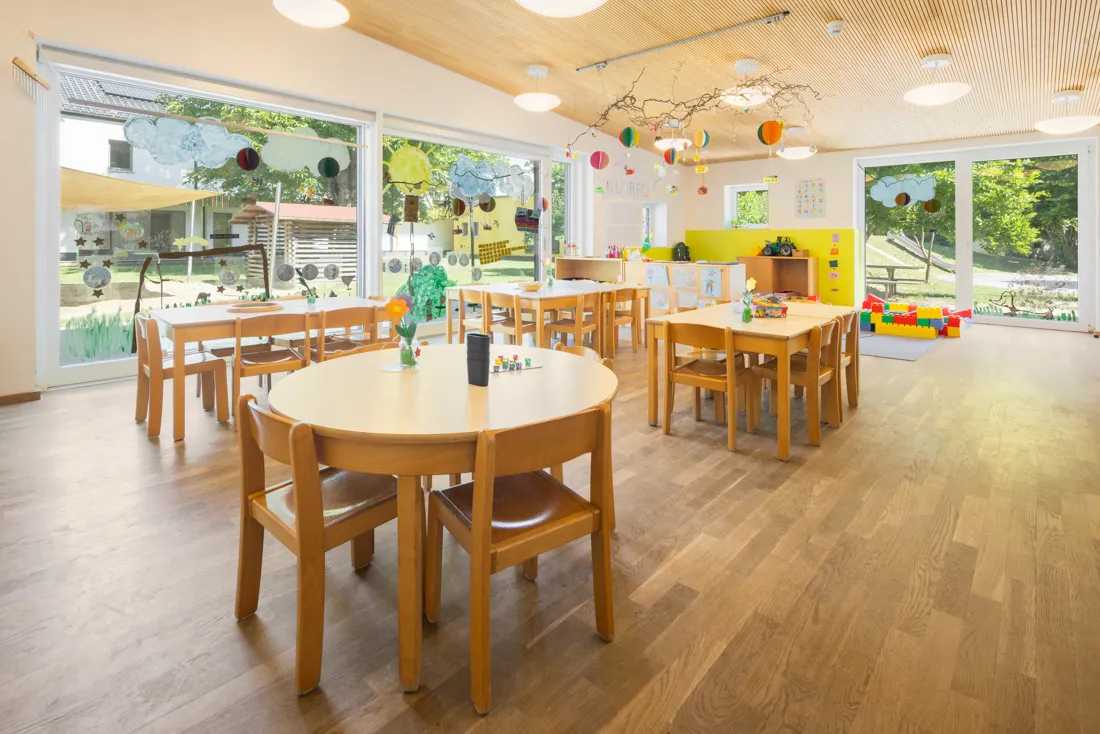 Gro&szlig;er offener Gruppenraum mit gro&szlig;en Fenstern und vielen Tischen und St&uuml;hlen zum Malen und Spielen